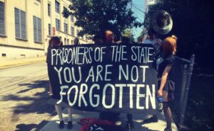 People outside a prison with a musical instruments and a banner reading "Prisoners of the state: You Are Not Forgotten"
