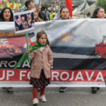 Small girl holding a sign featuring world leaders around a Kurdish boy with checmical burns, in front f a banner calling for protection for Rojava at a march