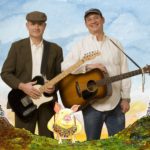 Promotional photo of Key Wilde (on the right) and Mr. Clarke (on the left). Mr. Clarke is wearing a brown suit & holding an electric guitar & Key Wilde is wearing a baseball cap & a white shirt & holding an acoustic guitar. Overlaid in front of them is a painting of a pig wearing many medals on its chest.