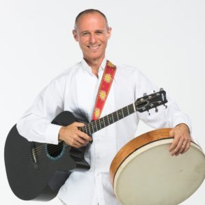 Image description: Billy Jonas wearing a longsleeved white shirt, smiling widely, and holding an acoustic guitar and a bodhran drum.